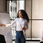 Young African American female coach or speaker make whiteboard presentation for diverse employees in office, focused biracial woman trainer present business project on flip chart at meeting